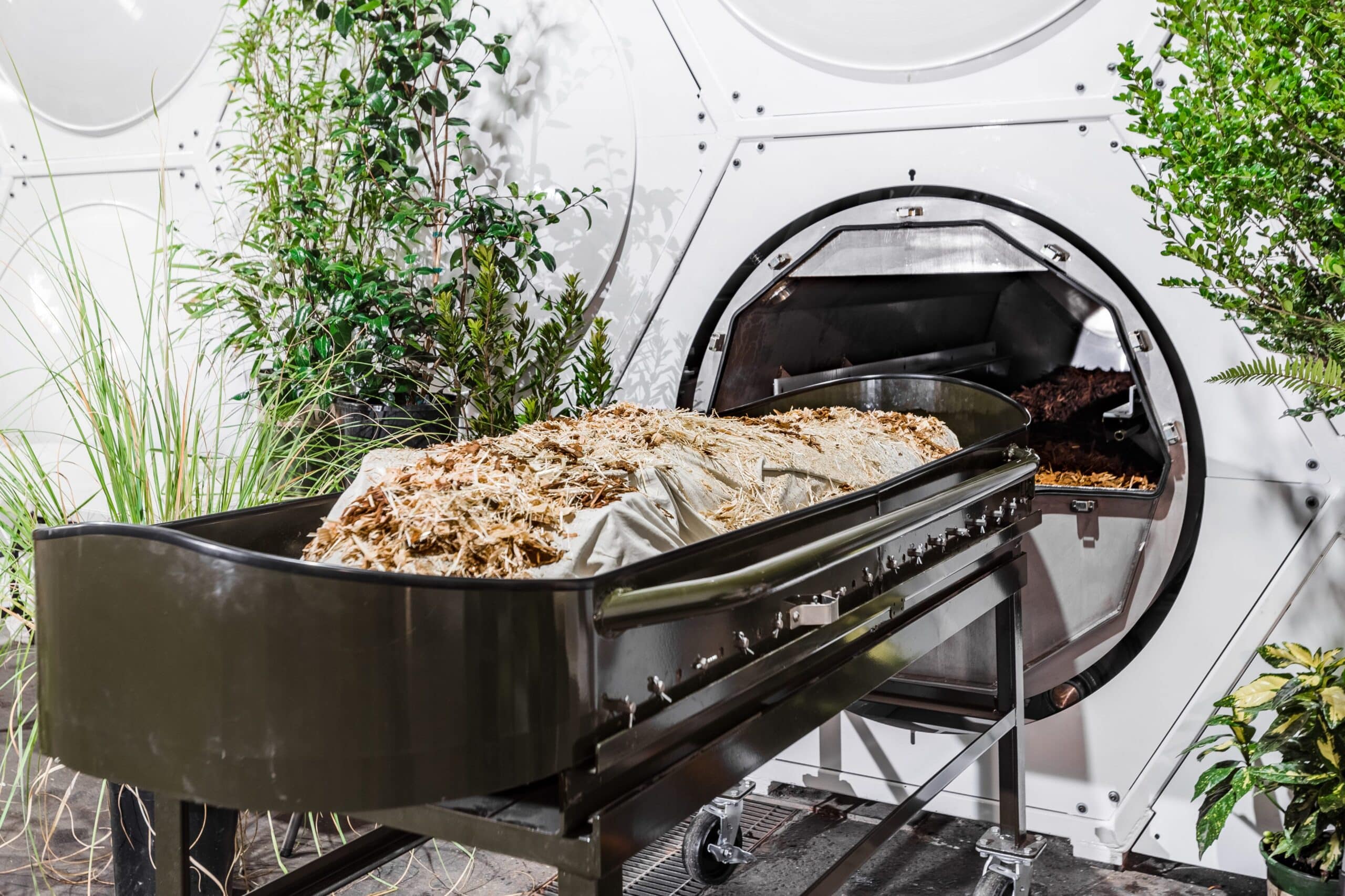 A dummy covered in plant material rests on a cradle in front of an open vessel. Each body is placed in a vessel with wood chips, alfalfa, and straw.