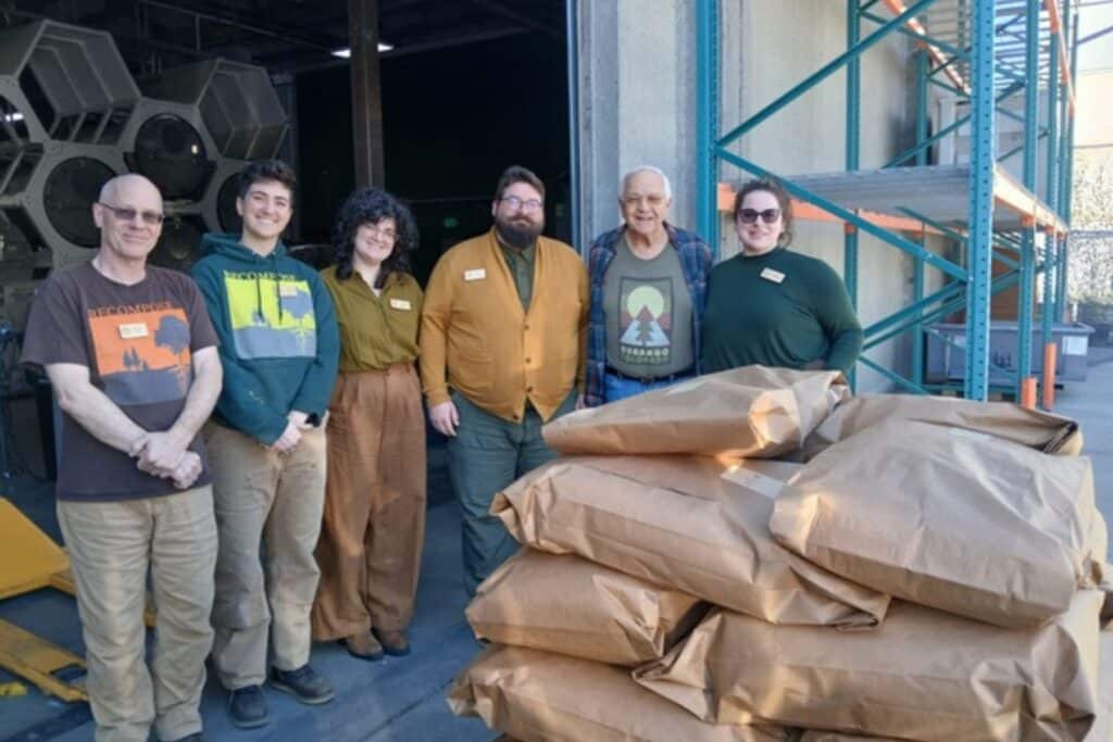 six people standing outside human composting facility with human composting bags piled up
